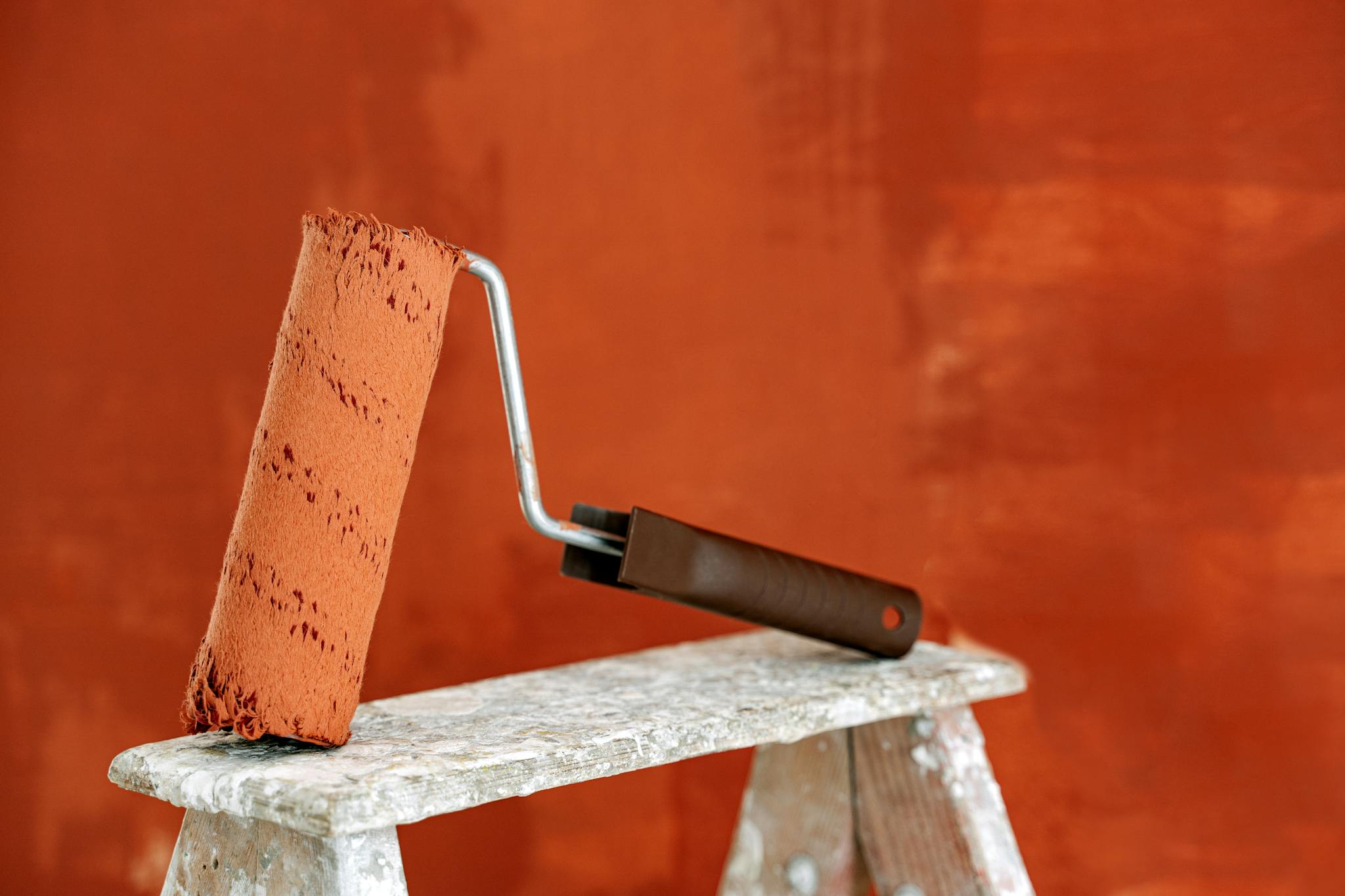 Detailed image of a paint roller resting on a ladder with a vibrant orange wall in the background.
